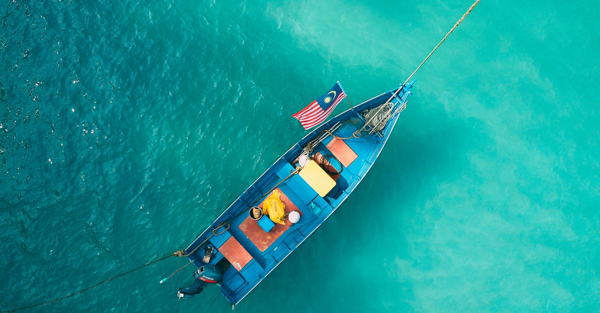 Drapeau de la Malaisie flottant à l’avant d’un bateau de pêche bleu, vu du ciel sur une mer turquoise